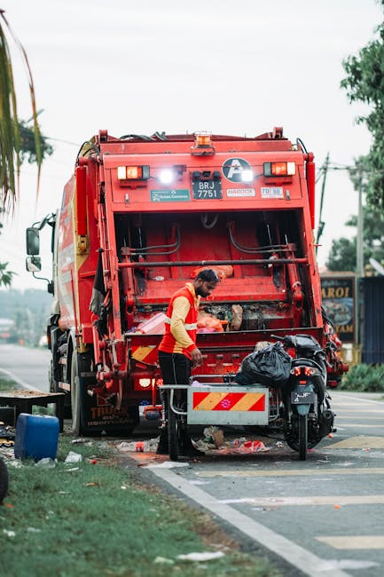 A waste collection worker dressed in a high-visibility vest and dark trousers is positioned behind a large red rubbish collection vehicle on a roadside. The vehicle's rear loading mechanism is open, revealing internal components and a small black trash bag resting on top of it. The worker appears to be handling or inspecting waste, with a black trash bag placed on the vehicle's platform nearby. The scene takes place on a paved street with a grassy verge and a blue plastic container on the ground in the foreground. In the background, there are trees, utility poles, and a few shops or buildings, suggesting an urban or suburban environment. The lighting indicates daytime, with natural light illuminating the scene, emphasizing the utilitarian purpose of the collection operation, which relates to private or alternative waste management services such as those offered by Waste Disposal Tooting.