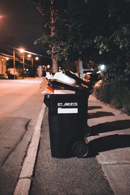 A waste collection worker dressed in a high-visibility vest and dark trousers is positioned behind a large red rubbish collection vehicle on a roadside. The vehicle's rear loading mechanism is open, revealing internal components and a small black trash bag resting on top of it. The worker appears to be handling or inspecting waste, with a black trash bag placed on the vehicle's platform nearby. The scene takes place on a paved street with a grassy verge and a blue plastic container on the ground in the foreground. In the background, there are trees, utility poles, and a few shops or buildings, suggesting an urban or suburban environment. The lighting indicates daytime, with natural light illuminating the scene, emphasizing the utilitarian purpose of the collection operation, which relates to private or alternative waste management services such as those offered by Waste Disposal Tooting.