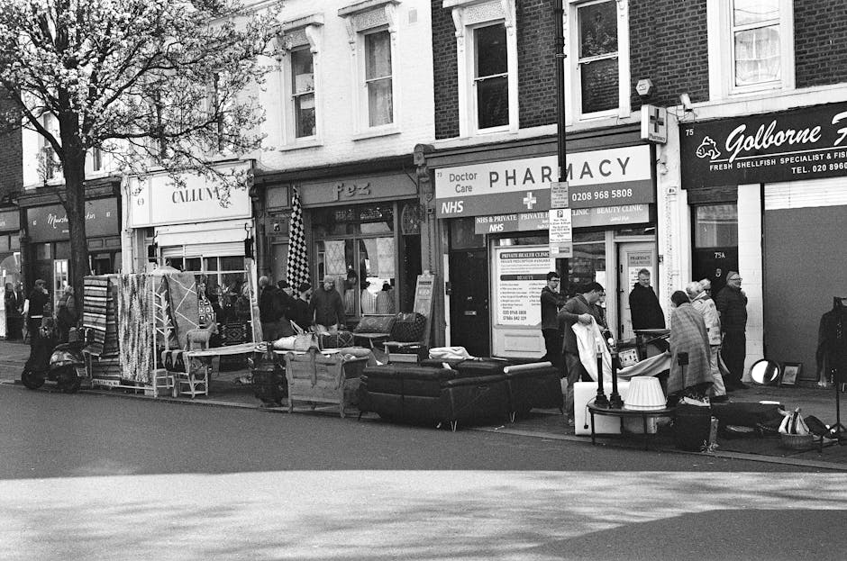 A busy street scene in front of a row of small shops and a pharmacy, featuring a variety of household and furniture items arranged along the sidewalk for sale or disposal. Visible items include a large black sofa, a smaller beige sofa or armchair, a desk lamp, and several stacked or leaning pieces of furniture such as chairs, tables, and shelving units. The furniture exhibits a range of textures, with upholstery fabric and painted or wooden finishes, primarily in dark and light tones. Several pedestrians, some looking at the items and others passing by, are gathered around the scene, with a few individuals inspecting or handling the objects. A delivery scooter is parked on the street to the left, and the background comprises storefronts, including a pharmacy with a prominent sign and a seafood specialist store with a smaller sign. The overall environment is illuminated by daylight, with natural shadows falling across the street and sidewalk, capturing an area where private or independent waste disposal activities take place in an urban setting, consistent with rubbish removal or on-site clearance services often provided by waste management specialists like Waste Disposal Tooting.
