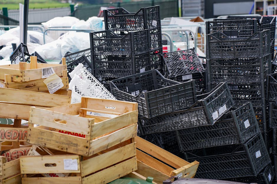 A large outdoor area filled with various types of discarded waste containers, including numerous black plastic crates stacked irregularly, some leaning and overlapping, with visible white labels on several. In the foreground, there are light-colored wooden crates, also stacked and containing miscellaneous packaging, with some labels and cardboard fragments visible. The background features additional white and transparent bags filled with waste or recyclables, with an industrial setting suggested by the fencing and metal structures. The scene appears well-lit with natural daylight, emphasizing the textures of the plastic, wood, and packaging materials. The overall environment suggests a waste management or rubbish collection site, where private refuse is gathered for disposal or alternative recycling methods, aligning with the services offered by Waste Disposal Tooting in terms of rubbish removal and onsite clearance solutions.