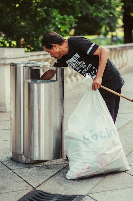 A middle-aged man wearing a black t-shirt and dark trousers is seen collecting litter from an outdoor stainless steel rubbish bin using his right hand, while holding a white plastic refuse bag in his left hand, which is partially filled with waste. The bin is cylindrical with a smooth, reflective surface and a top opening for waste disposal, positioned on a paved surface with visible concrete slabs. In the background, there are lush green trees and a stone balustrade, indicating a park or public space setting. The natural daylight highlights the urban environment and the clean, well-maintained appearance of the surroundings. This scene relates to waste management and rubbish collection, exemplifying on-site clearance or private waste handling, typical of waste disposal services in public areas, such as those provided by Waste Disposal Tooting.