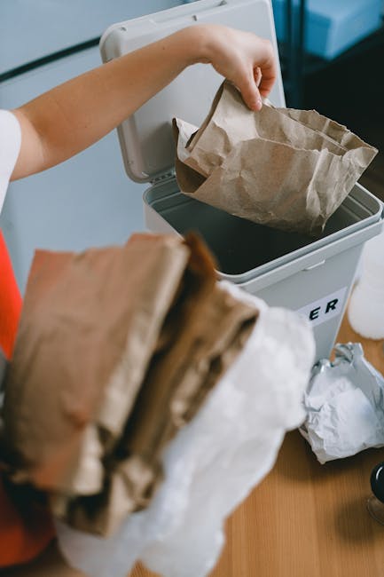 A person is disposing of crumpled paper bags into a grey trash bin, which has a partially visible label reading 'RUBBISH'. The bin is positioned indoors on a wooden surface, with a black interior lining visible at the top. The person's hand is gripping one of the paper bags, which appears to be made of brown kraft paper with a slightly textured surface. In the foreground, another crumpled paper bag is being held, out of focus, and partially obscuring the view of the bin. The environment suggests a domestic setting with natural lighting illuminating the scene, emphasizing the textures and earthy tones of the paper bags and the bin. This scene exemplifies an approach to private rubbish disposal or on-site waste collection, aligning with waste management practices that involve individual or household-level rubbish removal services, such as those provided by Waste Disposal Tooting.