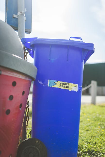 A large blue plastic wheeled rubbish bin with a closed lid, positioned outdoors on a grassy area next to a pink recycling container with black dots and a grey lid, both situated on a paved surface. The blue bin has a yellow sticker with the words 'Fight Back!' partially torn, and it appears to be in good condition with a textured surface. In the background, there is a metal pole supporting signage or fencing, and a clear sky suggesting a bright, sunny day. The scene is representative of private waste collection services, such as those offered by Waste Disposal Tooting, where individual rubbish bins are used for waste disposal and collection outside residential or commercial premises.